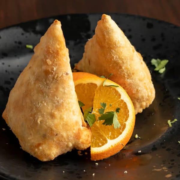 Two golden-brown Indian samosas with an orange slice and cilantro on a dark plate.