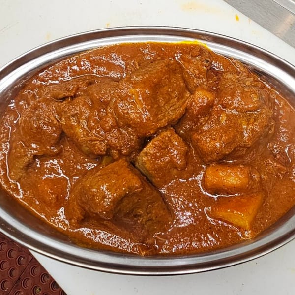 Close-up of a metal dish filled with rich, reddish-brown Indian curry and large pieces of meat.